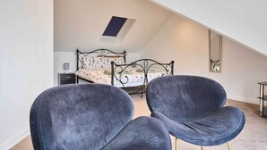 Un dormitorio con tragaluz, una cama con ropa de cama floral, dos sillas oscuras y una mesita de noche.