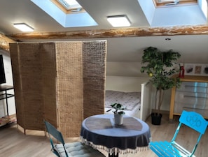 A room with a skylight, a bed, a dining table with chairs, a potted plant, and a kitchenette. - Studio in front of the Théâtre Molière | Mobility Pack (Sète)