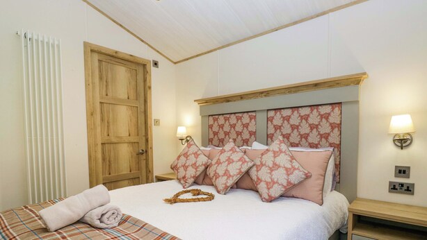 A bedroom with a bed, a wooden door, a bedside table with a lamp, and a wall socket. - Borwick Heights 17 (Carnforth)