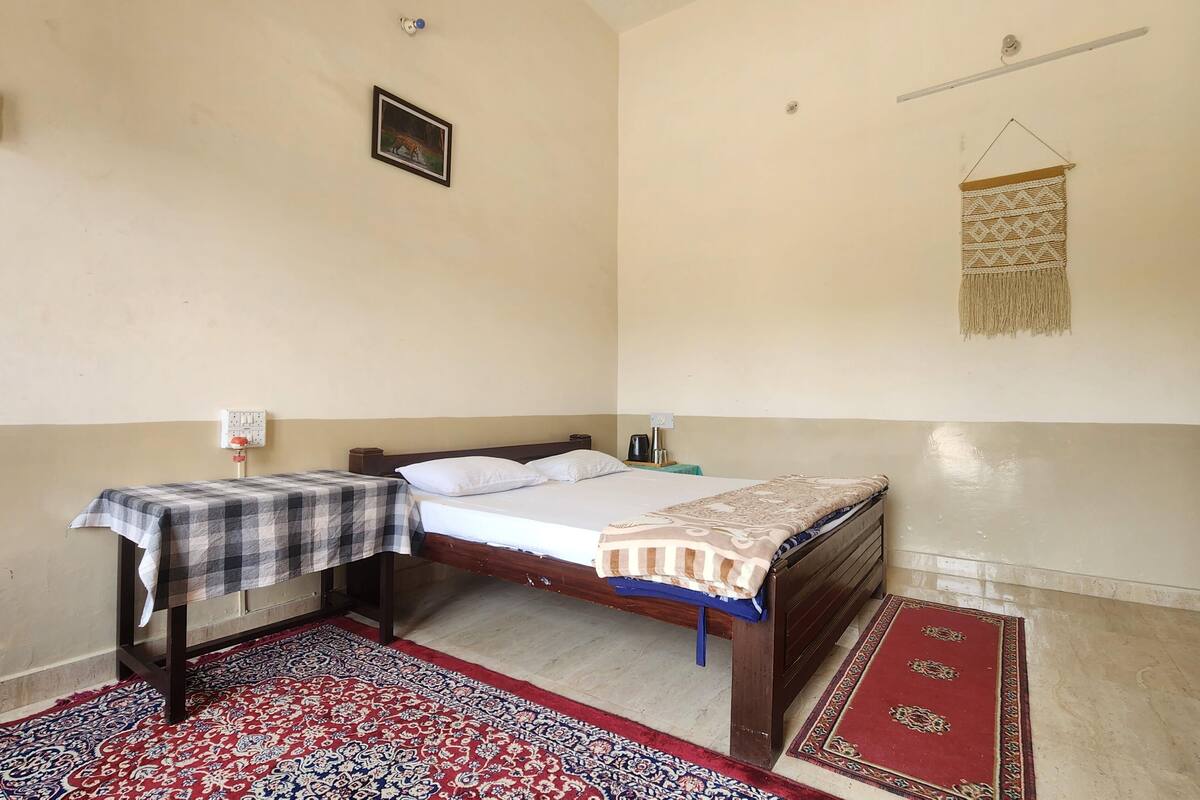 A bedroom with a bed, a nightstand, a table with a checkered tablecloth, a patterned rug, a wall-mounted light fixture, and a wall hanging.