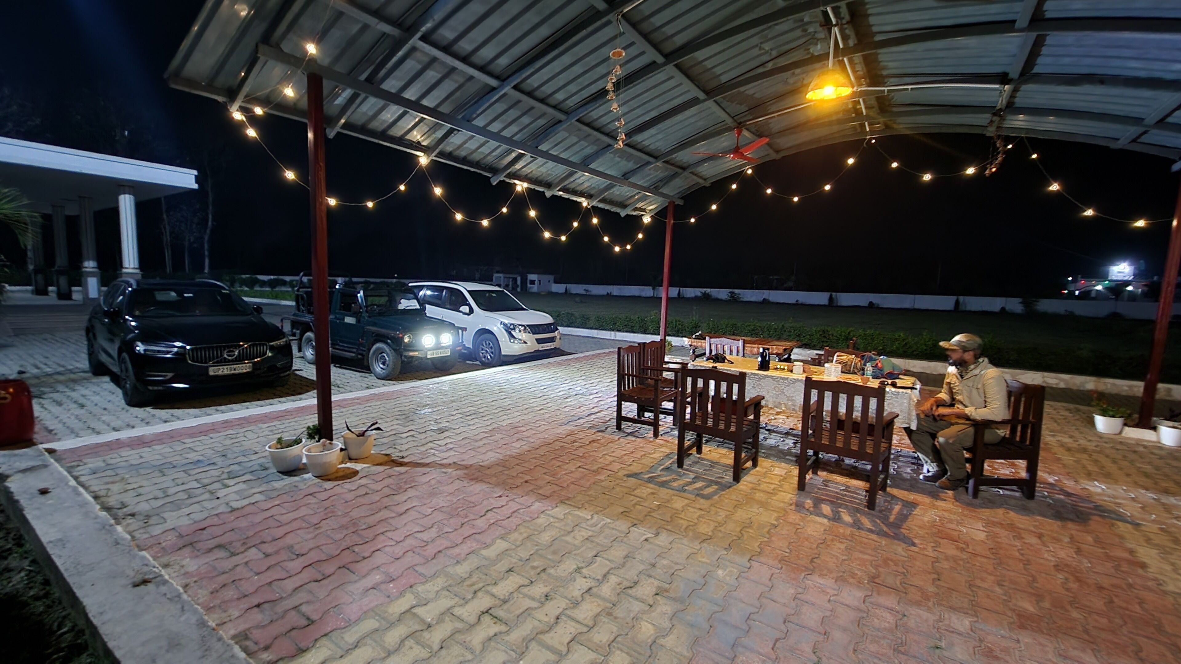 Une terrasse extérieure couverte, décorée de guirlandes lumineuses, avec une table et des chaises, et deux voitures garées.