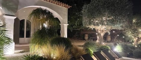 A covered outdoor seating area with lounge chairs, a pergola, and a well-lit garden at night.