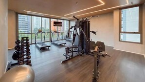 Un gimnasio con equipamiento de ejercicio, amplias ventanas y vistas a edificios.