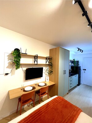 Una habitación compacta con cama, escritorio, dos sillas, televisor y kitchenette.