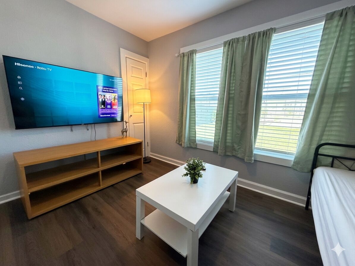 Ein Zimmer mit Flachbildfernseher, einem weißen Tisch, einer Bank, einem Bett und einem Fenster mit Vorhängen.