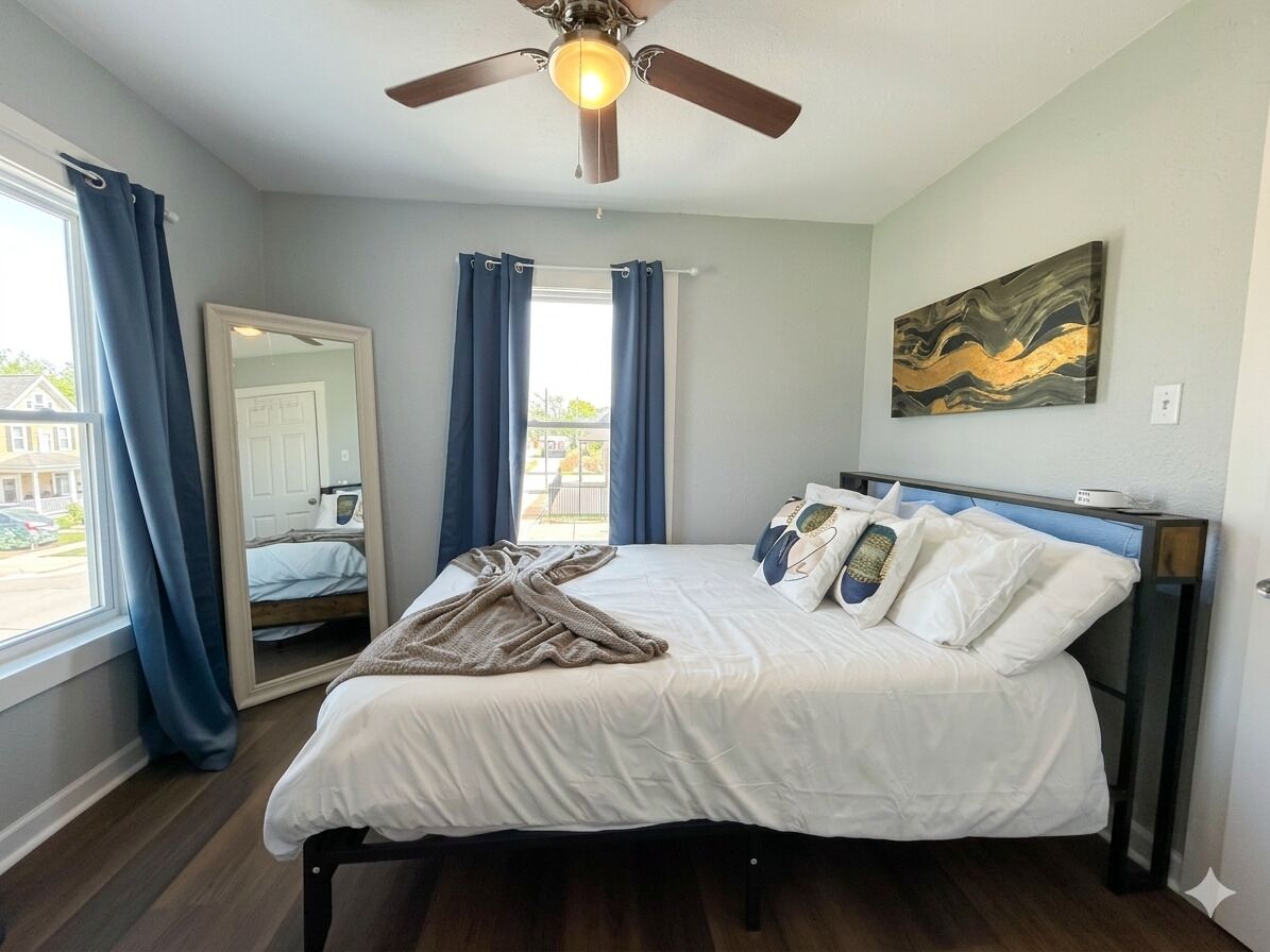 Un dormitorio con cama, ventilador de techo, ventana grande con cortinas, espejo y un cuadro en la pared.