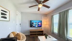 A living room with a ceiling fan, a flat-screen TV mounted on the wall, a white coffee table, a bean bag chair, and a window with curtains. - 2BDR+Extd Stay Discounts+Central+RemoteWork (Corpus Christi)