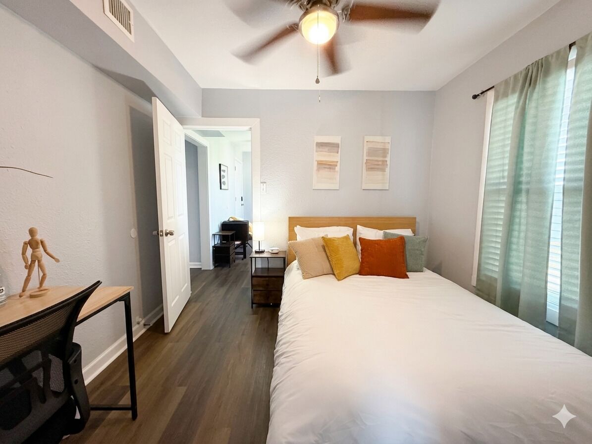 A bedroom with a bed, a desk, a chair, a ceiling fan, and a door leading to another room.