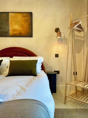 A neatly made bed with pillows, a bedside table, a wall-mounted light, and a wooden rack. - Fulham Design-led 2BR Apartment Near Olympia | Family Friendly | Sleeps 6 (London)