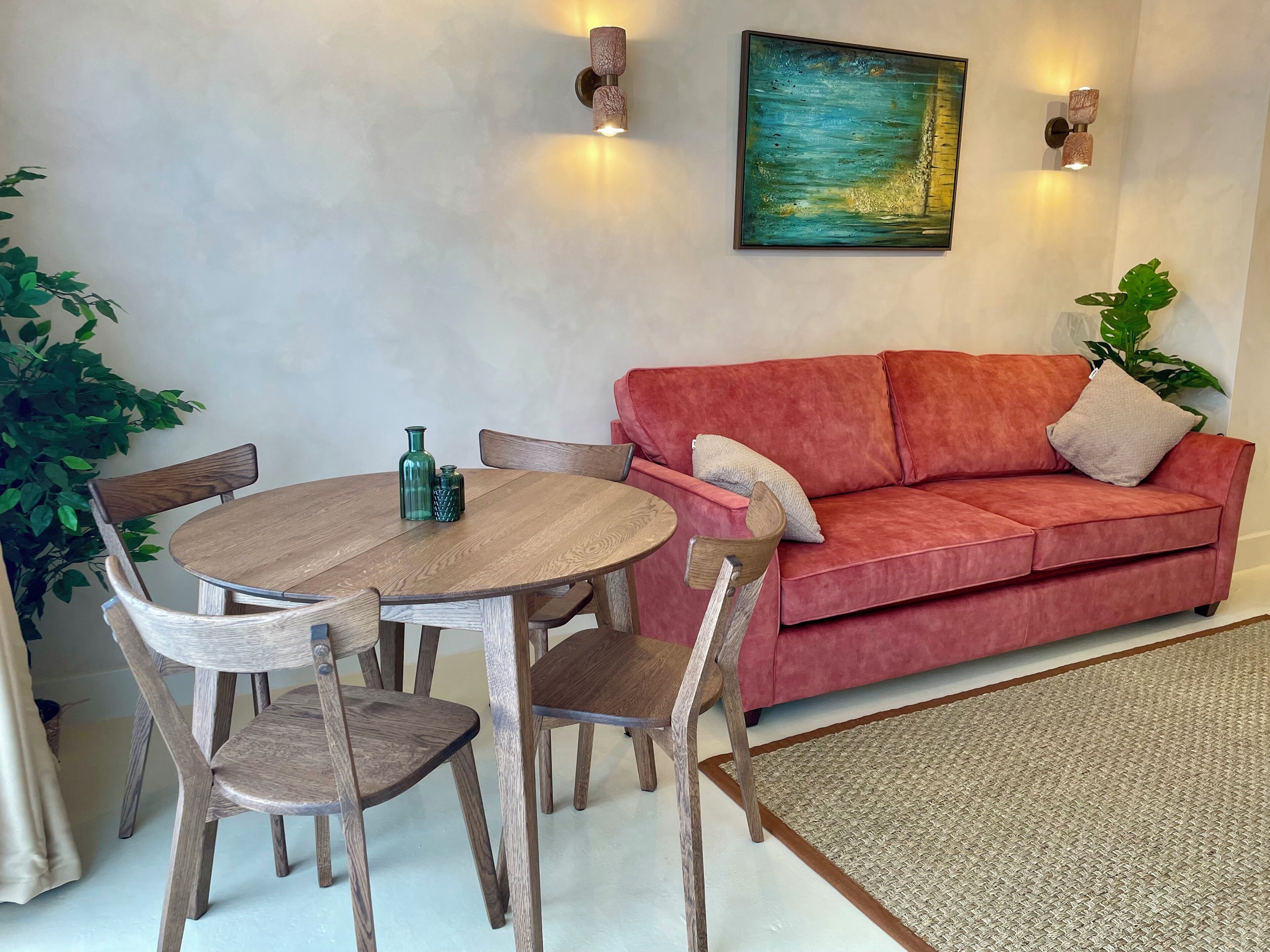 A living room with a round wooden table, four wooden chairs, a red sofa, two wall-mounted lights, a painting, and a potted plant.