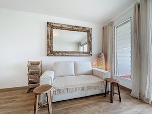 Ein Zimmer mit einer Couch, einem Holzschemel, einem Korbtisch, einem großen Spiegel und einem Fenster mit Vorhängen.