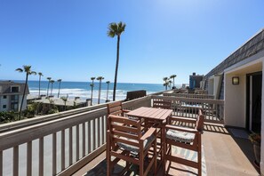 A balcony with wooden furniture, a view of the ocean, and palm trees.