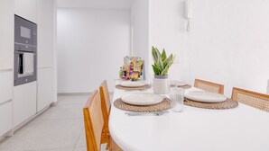 Una cocina moderna con armarios blancos, horno integrado y comedor con mesa blanca dispuesta con platos, cubiertos y una planta en maceta.