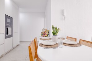 A modern kitchen with white cabinets, a built-in oven, and a dining area with a white table set with plates, cutlery, and a potted plant. - Casa Nubea • 500M from the beach • LVH (La Santa)