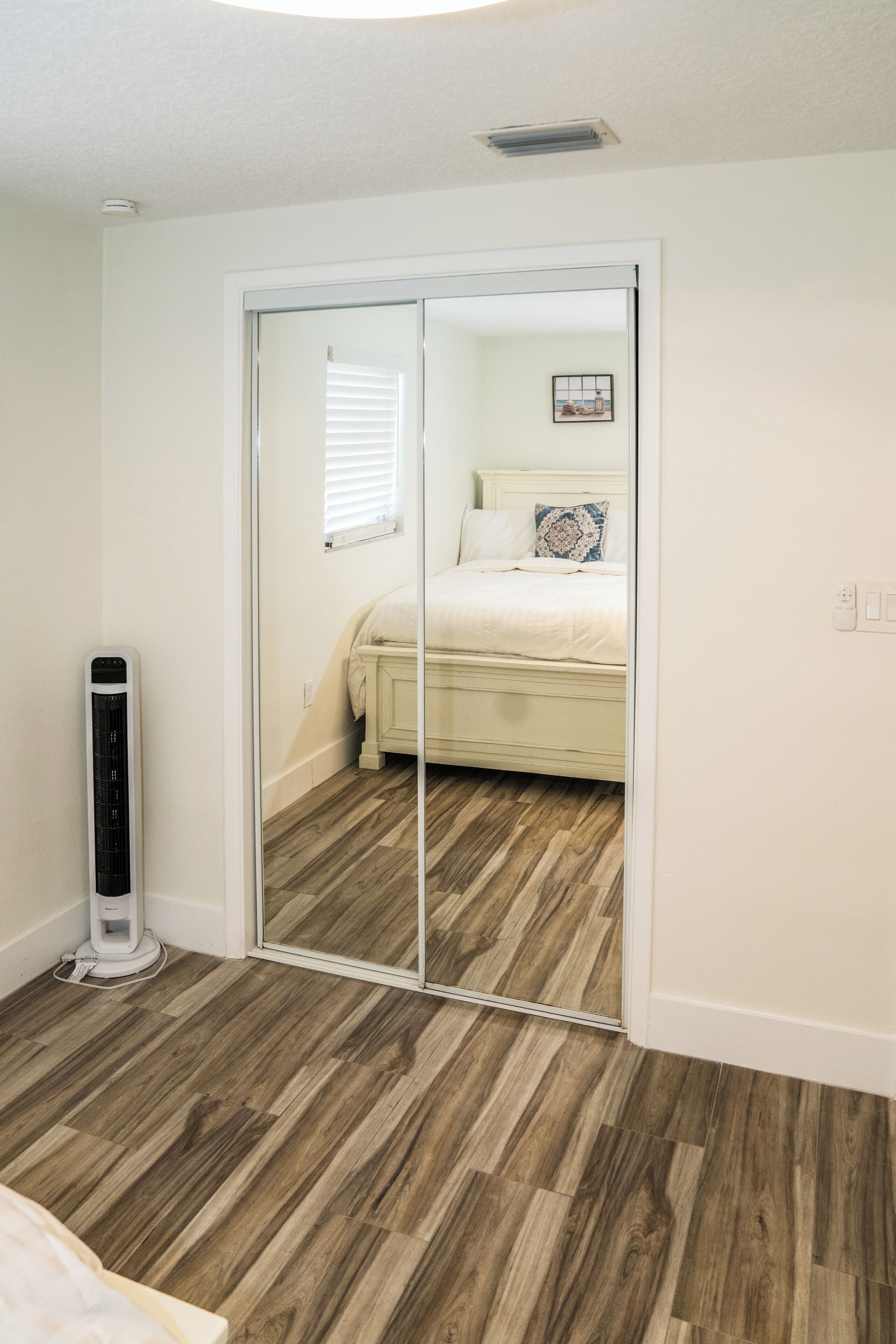 Un dormitorio con suelo de madera, una cama con ropa de cama blanca, una mesita de noche, un espejo y un calentador de pared.