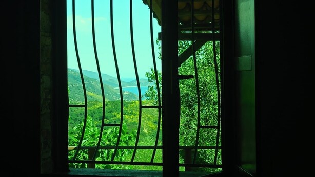Quarto com uma janela ampla com grades de metal, vista para colinas verdes e céu claro.
