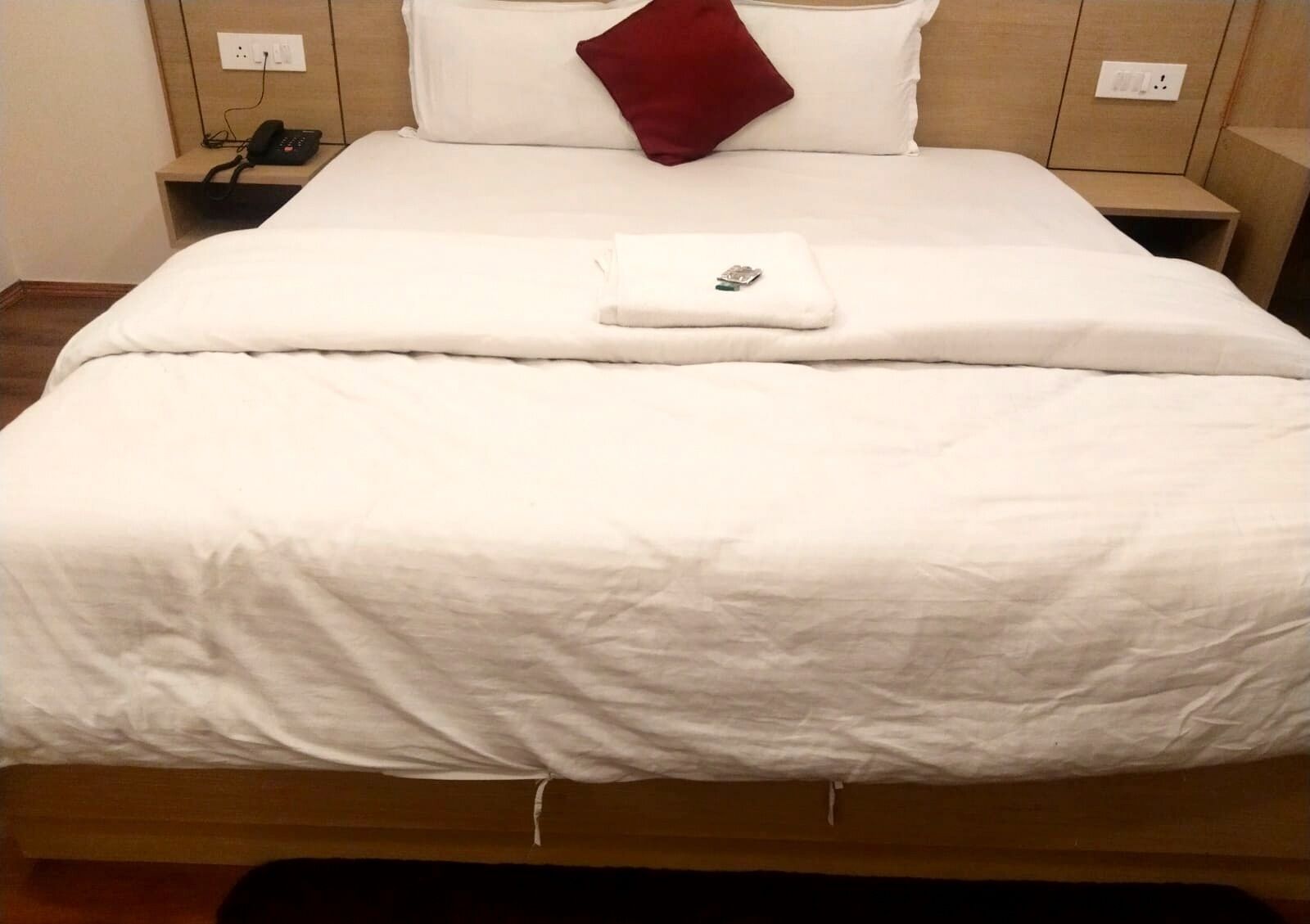 Una cama bien hecha con edredón blanco, almohada roja y una toalla doblada. Se observa una mesita de noche con un teléfono y un enchufe.