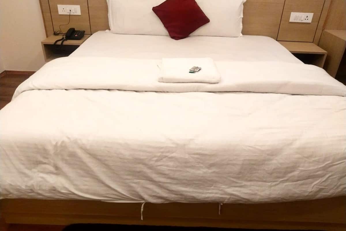 A neatly made bed with a white duvet, a red pillow, and a folded towel on the bed. There is a bedside table with a phone and a power outlet.