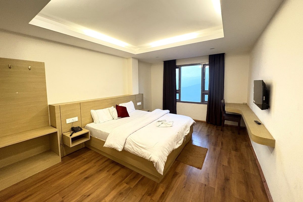 A hotel room with a wooden bed, a nightstand, a TV mounted on the wall, and a window with curtains.