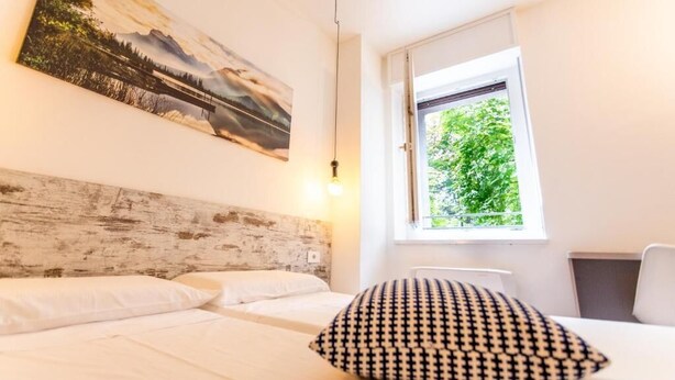 Une chambre à coucher avec une tête de lit en bois, un lit recouvert de draps blancs, un coussin à motifs, un tableau représentant un paysage et une fenêtre donnant sur un espace verdoyant.