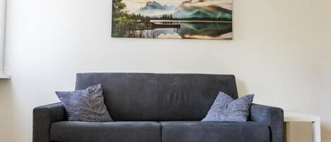 Un sofá gris con dos cojines estampados, una mesita de noche blanca y un cuadro de paisaje en la pared.