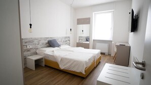A modern bedroom with a wooden floor, a bed with white bedding, a bedside table, a small stool, a wall-mounted TV, and a window with blinds.