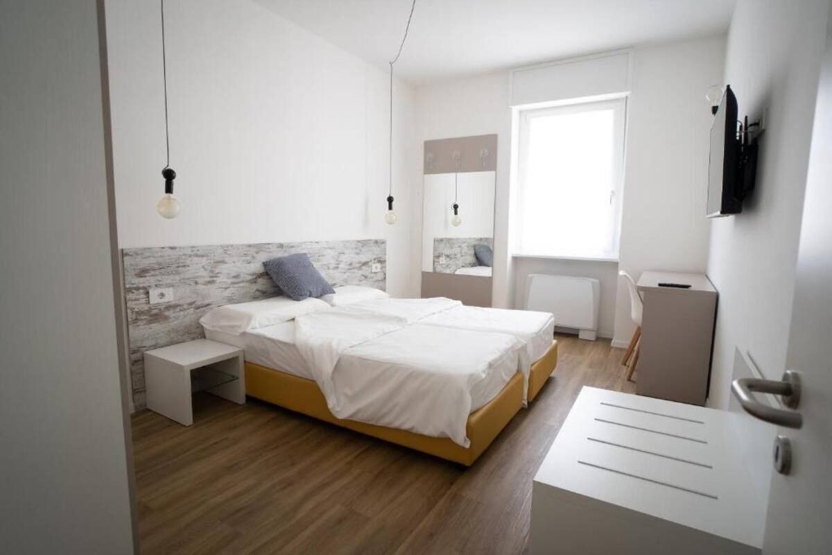 Quarto moderno com piso de madeira, cama com roupa de cama branca, mesa de cabeceira, um banquinho, TV fixada na parede e janela com persianas.