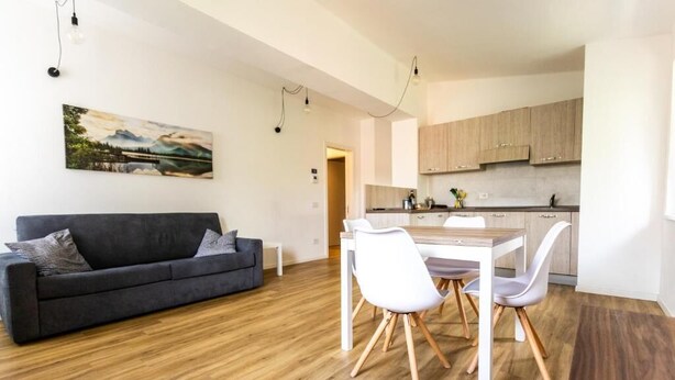 A modern living room with a dark grey sofa, white chairs, a wooden dining table, and a kitchen in the background.