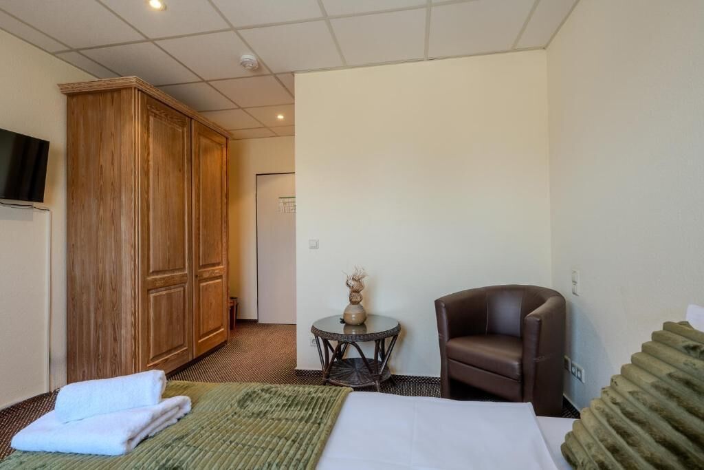Una habitación de hotel con una cama, un sillón de cuero marrón, una mesa redonda con un florero y un armario de madera.