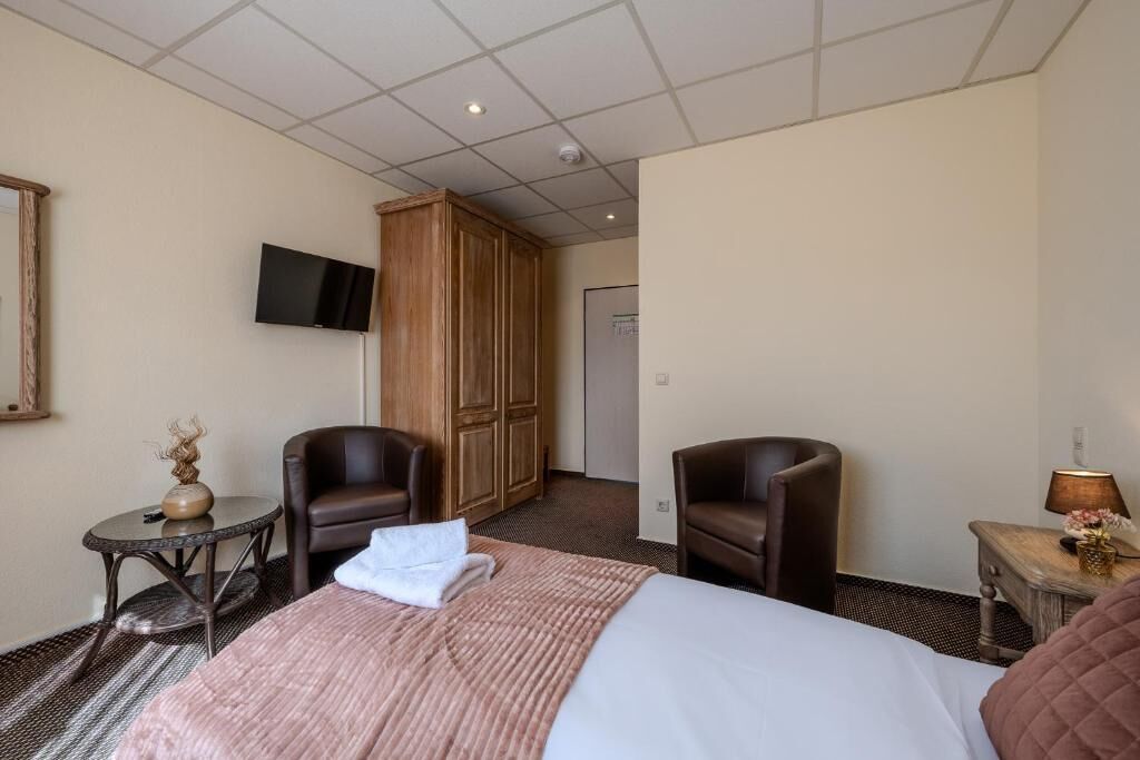A hotel room with a bed, two armchairs, a small table with a lamp, a television on the wall, and a wardrobe.