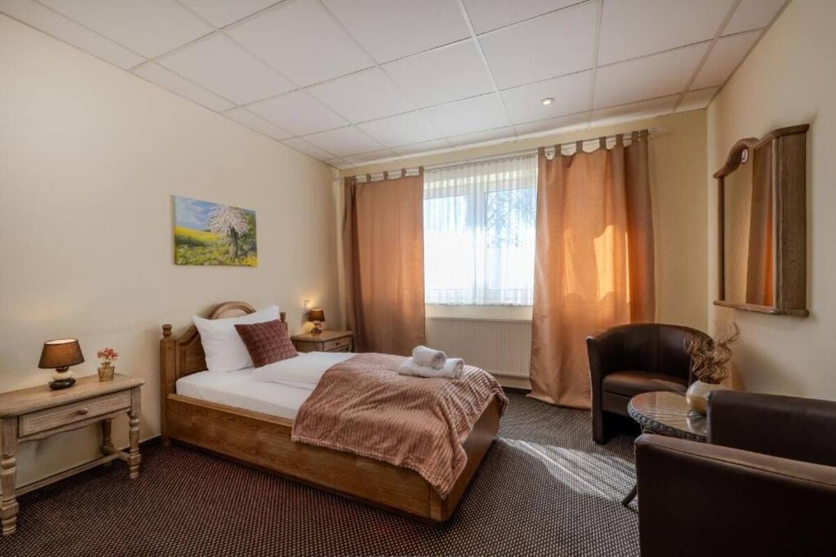 A hotel room with a bed, a chair, a small table, a nightstand, a painting, and a window with curtains.
