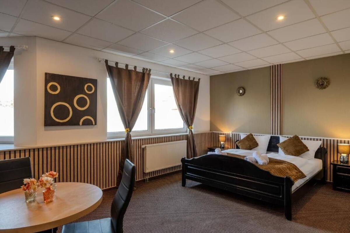 A hotel room with a bed, bedside tables, a round table with flowers, a radiator, and a window with curtains.