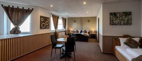 Habitación de hotel con cama, sofá, zona de comedor con sillas, mesa y ventana con cortinas.