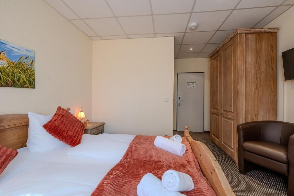 A hotel room with a bed, a wooden wardrobe, a brown leather chair, and a painting on the wall.