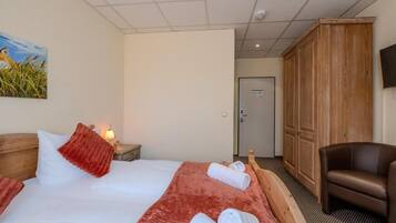 A hotel room with a bed, a wooden wardrobe, a brown leather chair, and a painting on the wall.