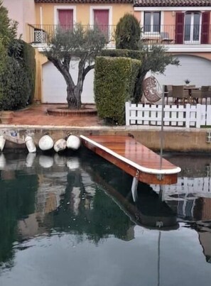 Eine Holzsteganlage erstreckt sich in ruhiges Wasser, umgeben von einem weißen Zaun mit Latten, mit einem Gebäude mit roten Fensterläden im Hintergrund.