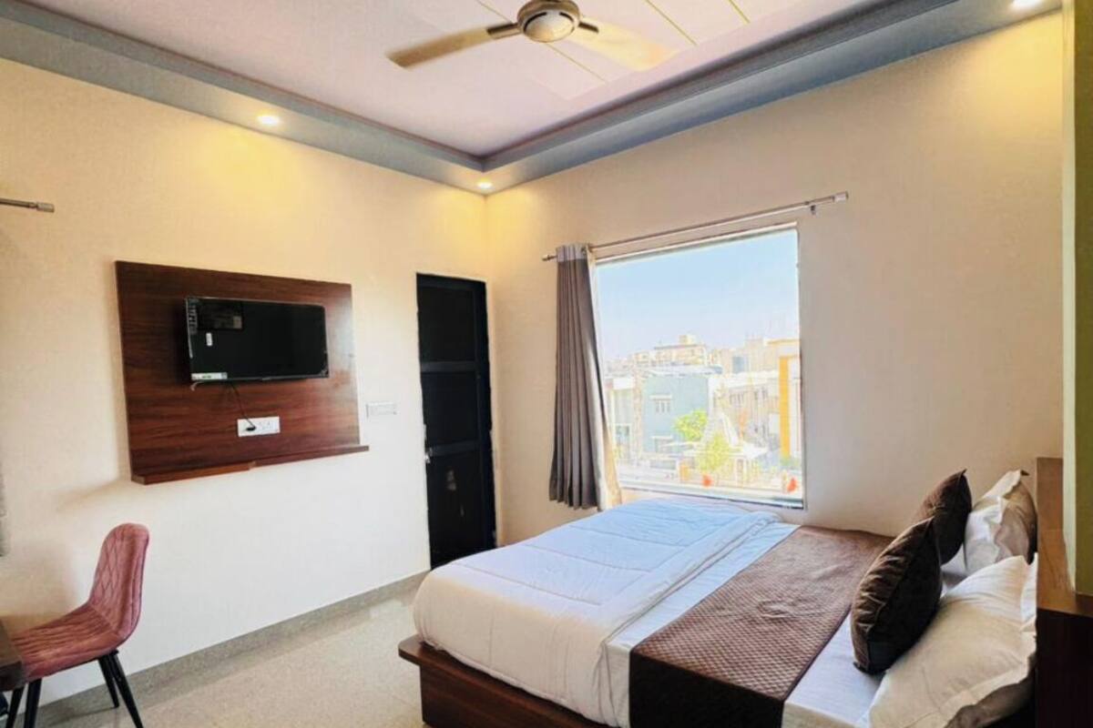 A bedroom with a bed, a TV mounted on the wall, a ceiling fan, and a window with a view of buildings.