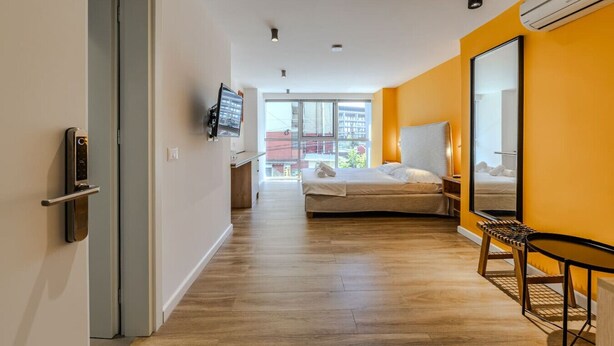 A modern hotel room with a bed, a desk, a chair, a TV, and a large window. - Orange Studio 104 | Love in Colours Collection by PikHost (Durrës)