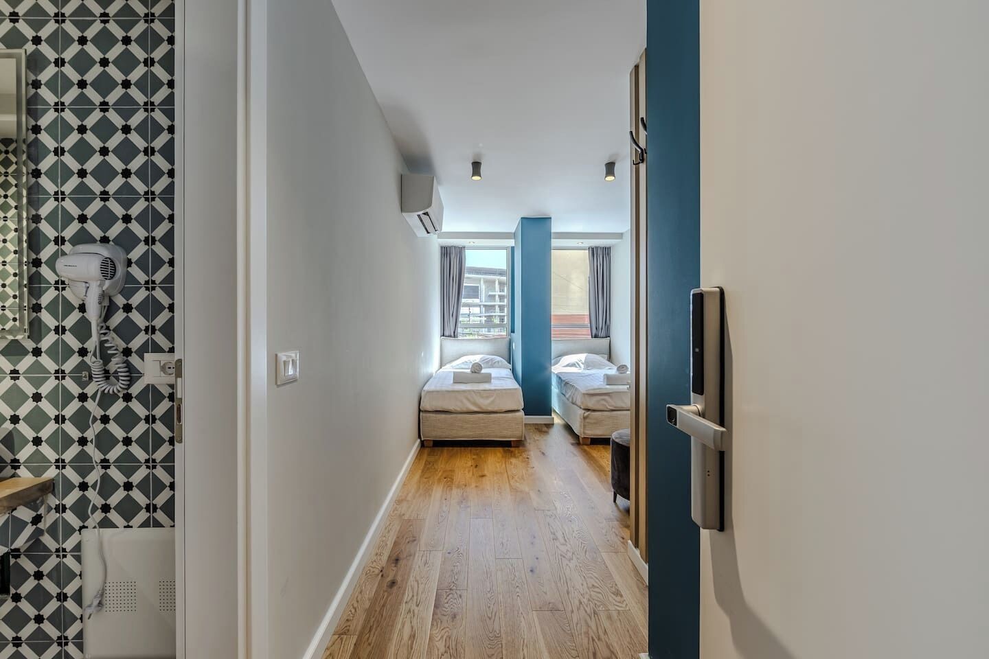 A narrow hallway leading to a bedroom with two beds, a hairdryer on the wall, and a window with curtains.