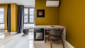 Quarto moderno com escrivaninha branca, cadeira cinza, banquinho redondo pequeno, obra de arte preta e janela com cortinas.