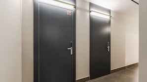 Duas portas pretas com os números 115 e 114, uma faixa de luz acima e chão de azulejos.