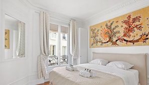 Ein Hotelzimmer mit Bett, großem Fenster mit Vorhängen, einem Gemälde an der Wand und einem Balkon mit schmiedeeisernem Geländer.