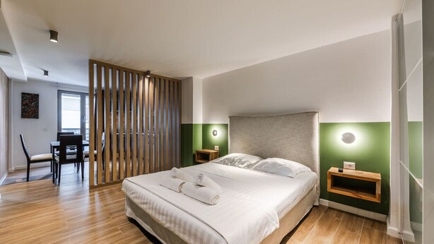 Ein modernes Schlafzimmer mit großem Bett, Holz-Kopfstütze, Nachttischen mit Lampen sowie einem Essbereich mit Stühlen und Tisch.