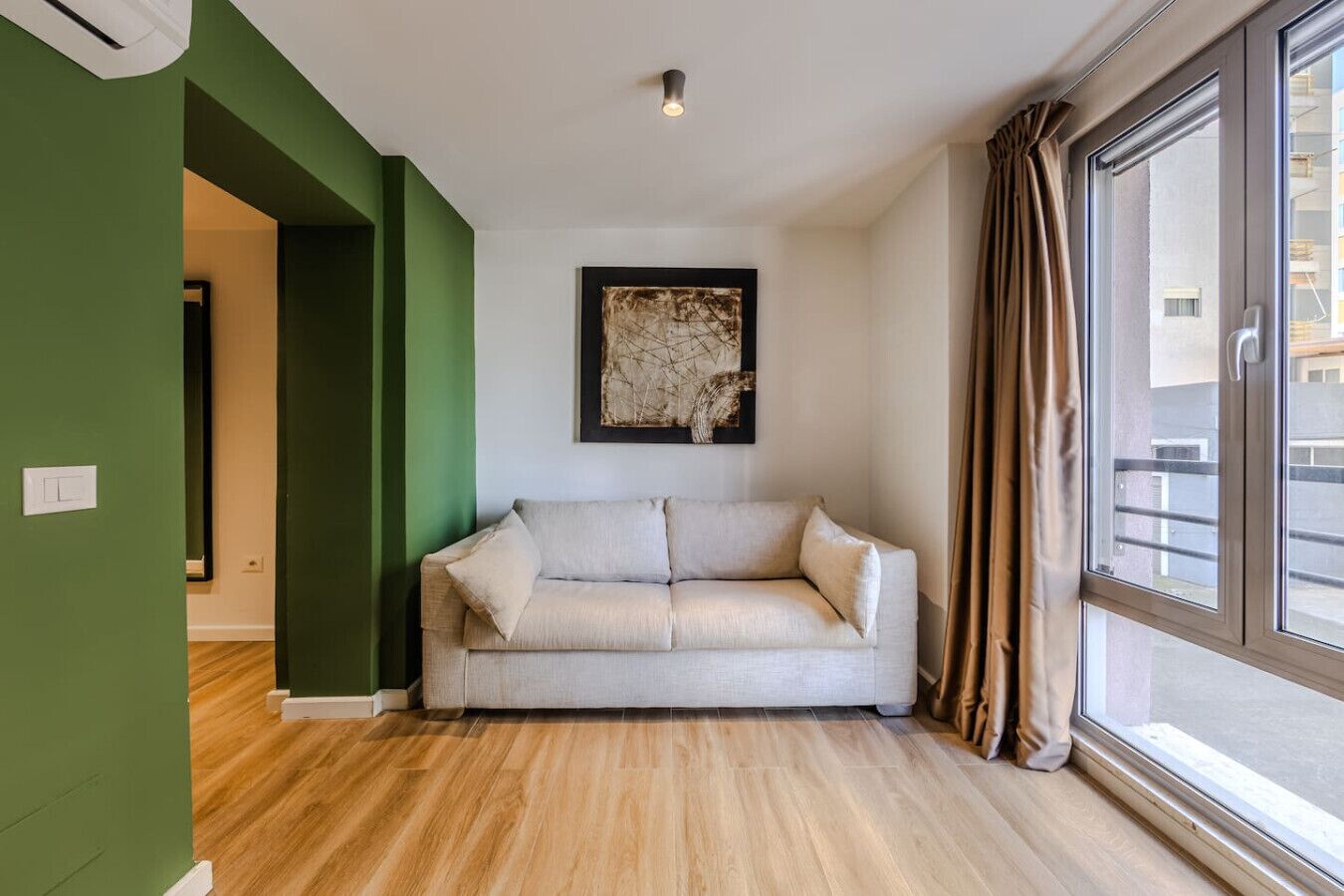 Ett rum med en beige soffa, ett inramat abstrakt konstverk, ett stort fönster med gardiner och en grön vägg.