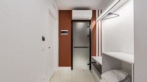 A narrow hallway with a door leading to a room, a wall-mounted air conditioner, and a bed with a white blanket. - Brown Studio 116 | Love in Colours Collection by PikHost (Durrës)