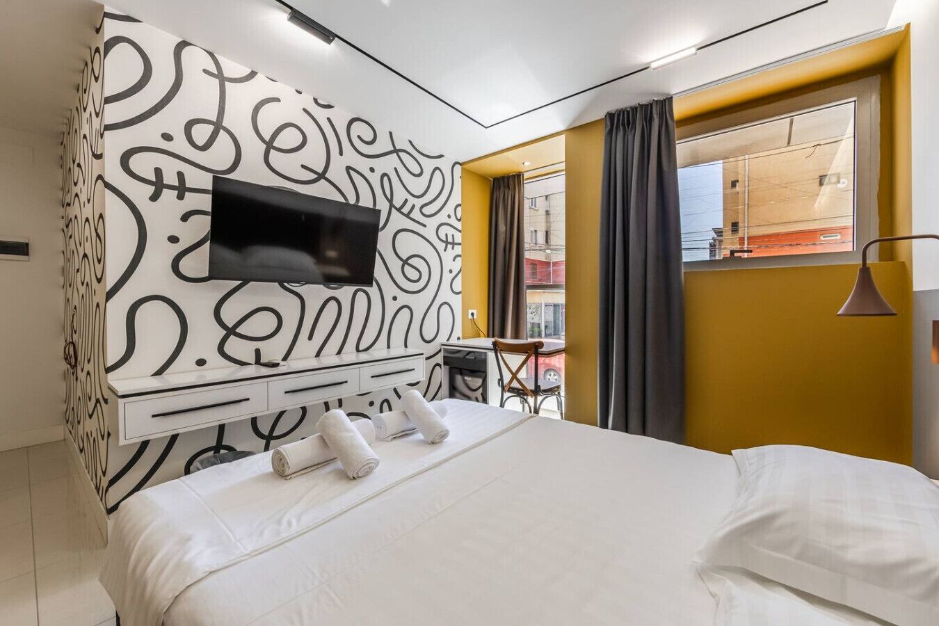 Hotellihuone, jossa on sänky, seinälle asennettu televisio, jonka ympärillä on koristeellinen kuvio, pieni pöytä ja tuoli sekä ikkuna, jossa on verhot.