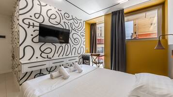 Hotellihuone, jossa on sänky, seinälle asennettu televisio, jonka ympärillä on koristeellinen kuvio, pieni pöytä ja tuoli sekä ikkuna, jossa on verhot.