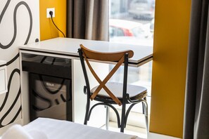 Habitación de hotel pequeña con un escritorio blanco, una silla de madera, una kitchenette compacta y una ventana con vistas a la calle.