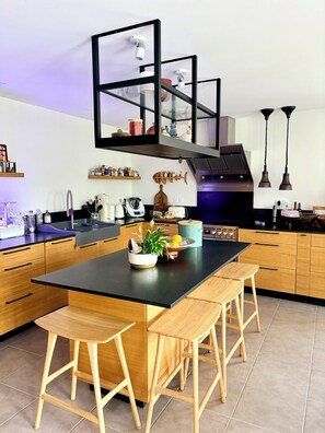 A modern kitchen with a central island, wooden stools, a fish-themed wall decoration, and a built-in oven. - Large charming house with enclosed garden near Rance, 15 min from St Malo and Dinan (La Vicomté-sur-Rance)