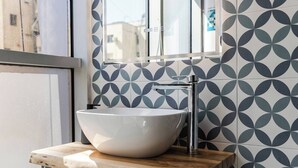 A bathroom with a white vessel sink, a chrome faucet, and a mirror above it. The wall has a geometric tile pattern. - Yellow Studio 106 | Love in Colours Collection by PikHost (Durrës)